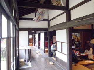 木枠の窓と白壁が続く古民家の廊下の右側にある畳の部屋で、人々が座って作業している写真