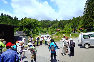 登山用の服装とポールを持った20人ほどの参加者が、山間の集落にある車が停まっている広場に集まっている写真