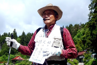 麦わら帽子をかぶった小野寺寛先生が、地名の謂れを書いた白い紙を胸に掲げ、ヘッドセットを使って話している写真
