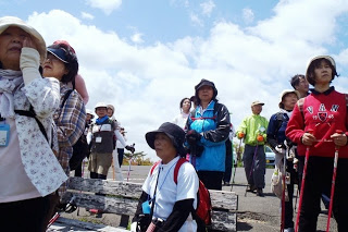 青空の下で帽子や登山用の服を身に着けた参加者たちが、眺めの良い風景を見ながらガイドの話を聞いている写真