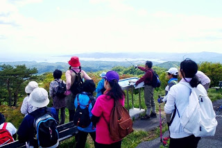 登山者たちが山の展望台から遠くの海と島を眺めており、ガイドがポールを向けて説明している写真