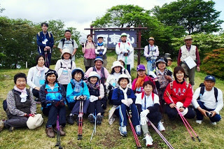 「田束山」と書かれた山頂の看板の前で、20人ほどの登山者たちが笑顔で並んでいる記念写真
