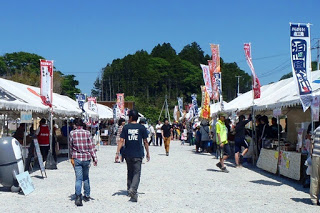 白いテントが両側に並ぶ屋外の会場で、多くの人が通りを歩き、色とりどりののぼり旗が風にはためいている晴天の中のイベント会場の写真