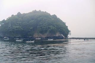 霧がかかった静かな海に緑の木々に覆われた小さな島が浮かび、その周囲を小型の漁船がゆっくりと進んでいる様子の写真