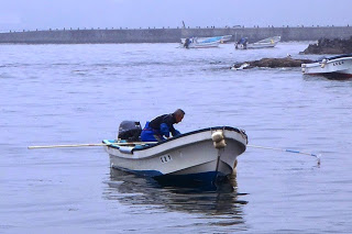 沖合で一人の漁師が小さなボートに乗り、身をかがめて海の中を覗き込むように作業している様子の写真