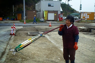 周囲で工事が行われている近くに立つ漁師が、長い柄の先にカギのような金具が付いた数本の道具を肩に担いでいる写真