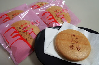 「最愛」という文字とモアイをモチーフにして描かれたピンク色の個包装に入った丸い最中と皿の上の白いナプキンにのっている「最愛(もあい)」のお菓子を写した写真