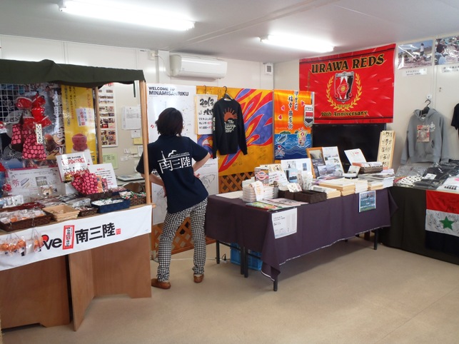 手作りの小物や地元のグッズなどが机や棚に並び、壁には浦和レッズの大きな赤い旗が飾られた、店内で、南三陸町のTシャツを着た女性店員が腰に手を当ててポーズをとり後ろ姿で写っている写真