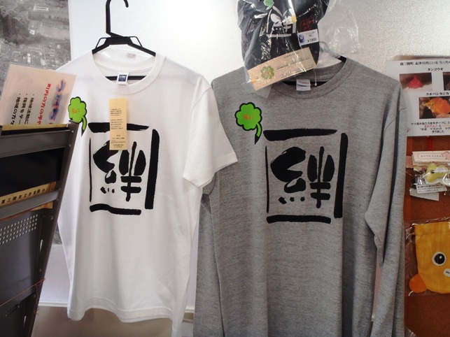 中央に「絆」という大きな漢字が黒く印刷されていて、左胸には緑色の四つ葉がデザインされた白とグレーの2色のTシャツが壁にかけて陳列されている写真