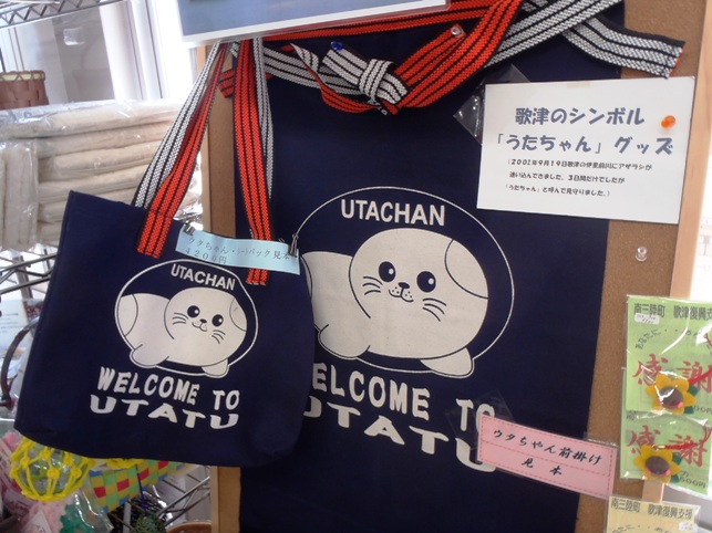 「UTACHAN」と書かれた白いアザラシのキャラクター「うたちゃん」が大きく描かれた紺色のトートバッグと前掛けが展示されており、横に「歌津のシンボル『うたちゃん』グッズ」と書かれた紹介文も貼られているうたちゃんグッズの写真