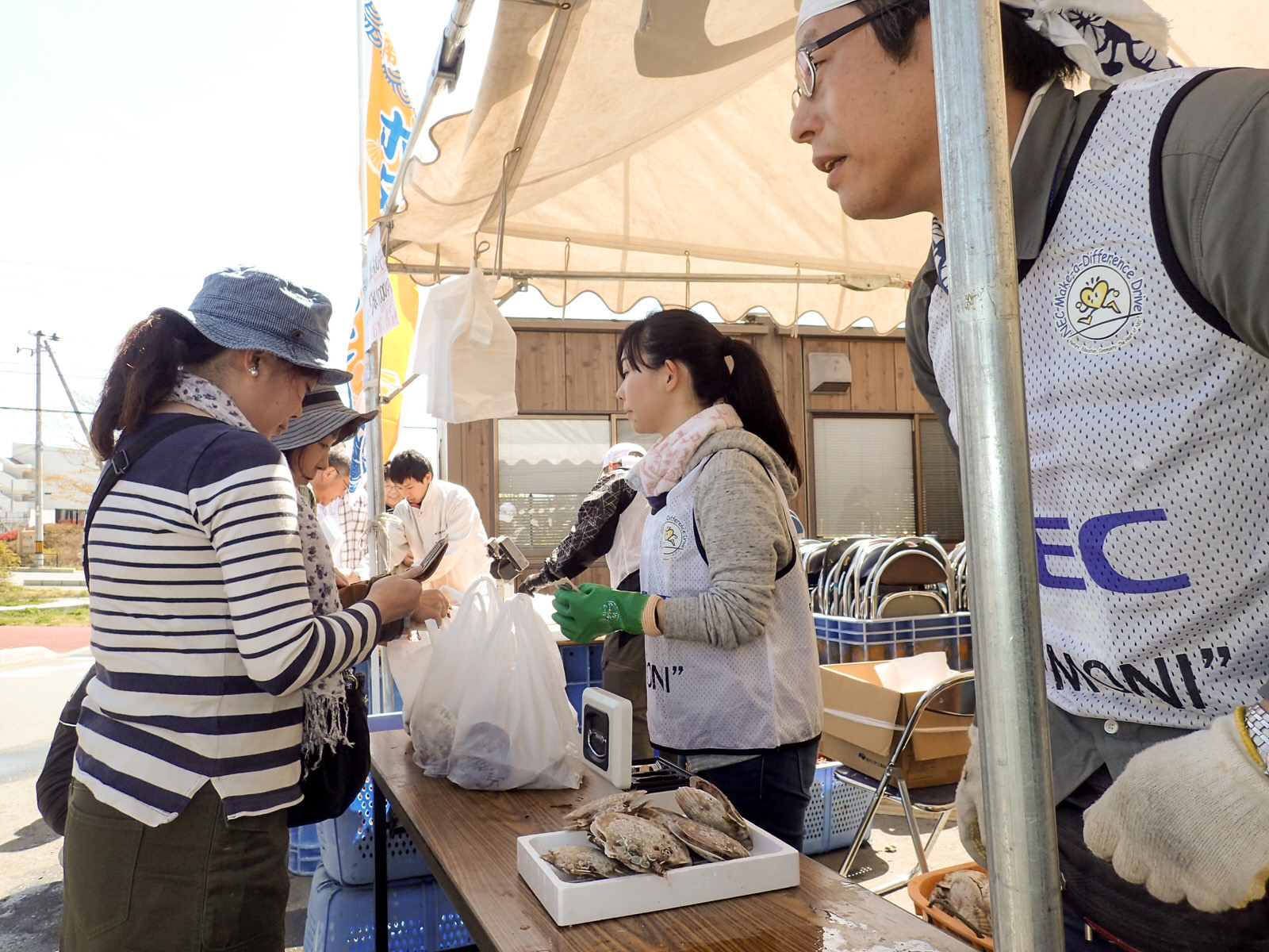 出店のテントで、ボランティアのスタッフが新鮮な貝類を並べて来場者と笑顔でやりとりしながら販売している写真