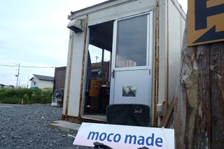 錆びた金属製のコンテナの前に「moco made」と書かれた白い看板が立てかけられている店舗の外観を写している写真