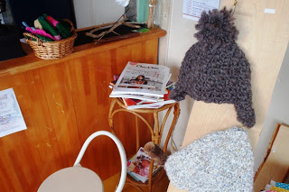 木製のカウンターの上に編み物が入ったバスケットが置かれており、その隣には雑誌や新聞が積まれた小さなテーブルと白い椅子があり、壁には毛糸で編まれた帽子が掛けられている様子が写っている写真