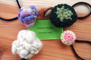 異なる色と模様で編まれた手作りの飾りが付いたヘアゴムが4つ並べられており、中央には「moco made」と「tomoko yano」と書かれた緑色のカードが置かれている写真