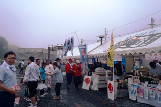 白いテントの屋台の前に多くの来場者が並び、地元の特産品や食べ物を買い求めている様子を霧がかった空の下で撮影した、南三陸夏まつりの出店エリアの写真