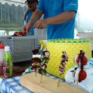 トマトやタコ、ピーマン、などを串に刺して焼いている男性たちの手元と、串焼きが並べられた木の台が映っている、屋台で準備される地元食材の串焼きの写真