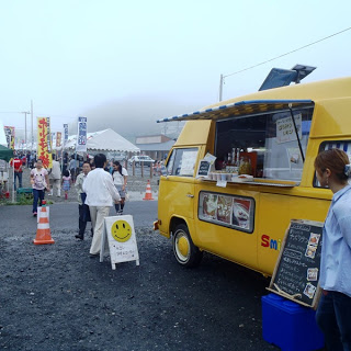 黄色いフォルクスワーゲンのキッチンカーの前で人々が飲み物や軽食を購入しており、黒板メニューとスマイルマークの看板が置かれている、フェスタの移動販売車の写真