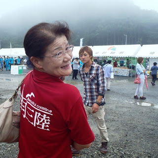「南三陸」と書かれた赤いTシャツを着た女性が笑顔で振り向き、祭り会場のテントや来場者が背景に広がっている祭り会場を写した写真