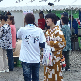 白地に「南三陸」と書かれたTシャツを着た女性と、茶色の浴衣を着た女性が談笑している、観客とボランティアスタッフが交流する南三陸夏まつりの会場の一角の写真