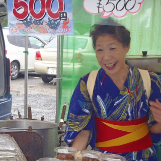 青と黄色の浴衣に赤い帯を締めた笑顔の女性が屋台で販売している様子の写真