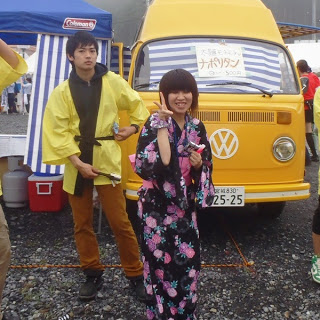 黄色の法被を着た男性と、黒地にピンクの花柄の浴衣を着てピースサインをしている女性が、黄色のフォルクスワーゲンの前で写っている写真