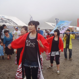 赤い法被を羽織った若い女性たちが笑顔で踊りながら会場内を練り歩き、周囲の観客がそれを見守っているコンテストの様子の写真