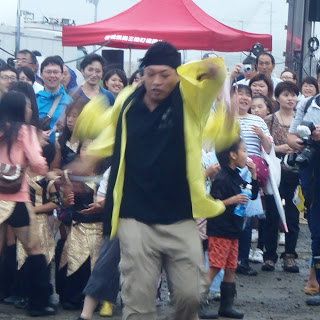 黄色い法被を着た男性が躍動的な動きで踊りながらパフォーマンスを披露し、後ろには拍手を送る子どもや大人が楽しそうに見守っている様子の写真