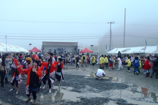 雨上がりで地面に水たまりが残る中、赤や黄色の衣装を着た踊り手たちがステージ前に集まり、観客も巻き込んで盛り上がっている様子の写真