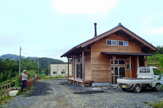 木造の平屋建ての小さな家の前に軽トラックが停まり、横には男性がフェンス沿いに立っている山間の風景の写真