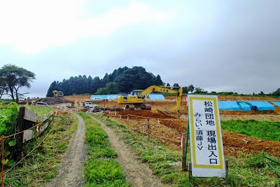 未舗装の道の先に松崎団地の現場出入口を示す看板が立ち、重機が造成工事を行っている様子が見える写真