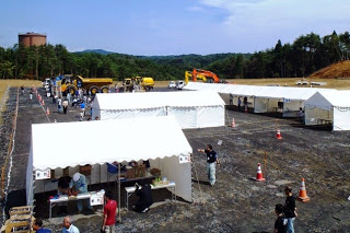 木々に囲まれた平坦な広場に白いテントがいくつも並び、その周囲でスタッフや来場者が活動しており、奥には重機が数台並んでいる見学会の全景写真