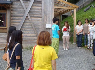 木造の建物の前で、Circle Japanのメンバー4名とYes工房の皆さんが向かい合って立ち、1人の女性がオクトパス君のぬいぐるみを手に持ち見つめている写真