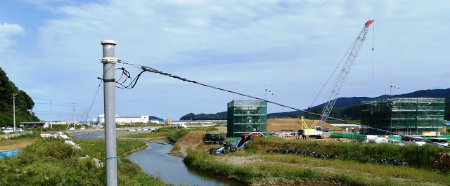 川の手前に電柱が立ち、川沿いに足場の組まれた建設中の橋脚やクレーン車や重機が写された写真
