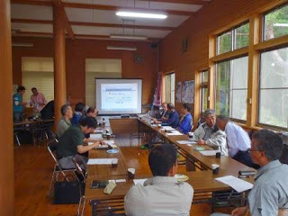 大きな窓から自然光が差し込む中、長机をコの字型に並べた会議室の前方に資料が映し出されたスクリーンが設置され、参加者が資料を見ながら内容を確認しているセミナーの様子の写真