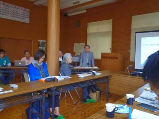 文字が映されているスクリーンが室内前方に設置され、左側前方に立って話す男性の周りで、参加者が座って真剣に話を聞いている写真