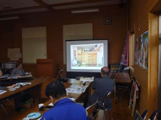 照明を消して薄暗くなった室内に設置されたスクリーンに木造の室内写真が映され、参加者がそれを見ながら資料を確認している写真