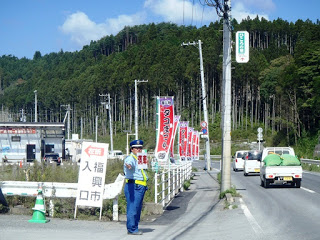 青空の下、山林を背景に「福興市入口」と書かれた看板前で、交通整理員が手で一時停止の指示を出し、交通整理をしている様子の写真