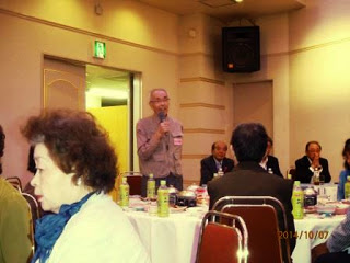 円卓に料理と飲み物が並び、複数の人が着席する中、一人の男性が立ってマイクを持ち、挨拶をしている様子の写真