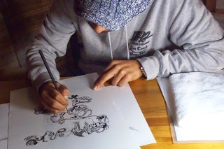 グレー色のパーカーを着た男性が、机に向かってペンで白い紙にイラストを描いている様子の写真