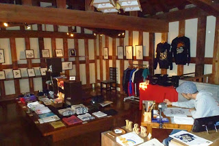 木の柱が並ぶ落ち着いた色合いの店内に、Tシャツやアート作品、雑貨などが並び、右側の机で男性が一人作業している様子の写真