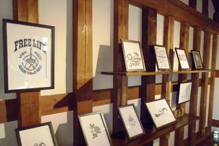 木の壁に設けられた棚に、「FREE LIFE」などの文字が描かれたイラスト入りの額縁が等間隔に並んでいる展示スペースの写真