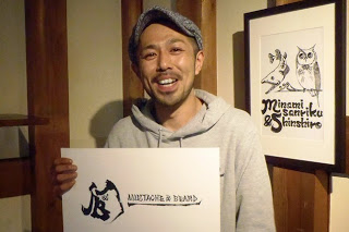 壁にフクロウのイラストが飾られている店内で、灰色のパーカーと帽子をかぶった男性が笑顔で英字で店名が書かれた作品を持っている写真