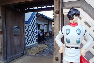 レトロな木造建物の門の横に、白い服を着たアニメ「サイボーグ009」のキャラクターの人形が立っている施設の入口の写真