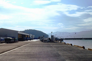 港の岸壁に沿って倉庫やトラックが並び、遠くの山と漁船が静かな海に浮かんでいる様子が朝の光に照らされている、静かな漁港の写真