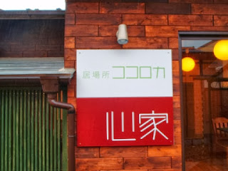 木目調の外壁に「居場所ココロカ」と「心家」と書かれた看板が掲示されている写真