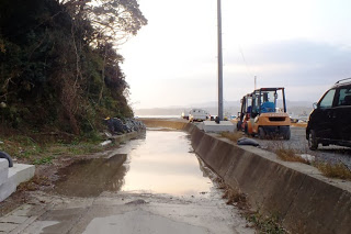 右側には工事用の重機と車が並んで停まっており、左側には低くなり水溜りが続く道が続いている写真