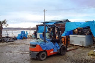 海のそばの作業場に赤と黒のフォークリフトが停まっており、周囲には青いビニールシートや漁具などが置かれている様子の写真
