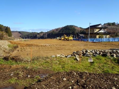 重機が複数台置かれた整地された広い造成地の隣に、すでに建っている一軒家が見える西戸地区の災害公営住宅予定地の写真