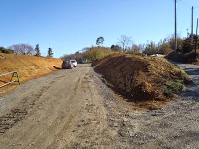 左右に盛土された道の中央に砂利が敷かれた仮設道路が続き、工事車両が奥に停まっている造成地内の道路整備中の様子の写真