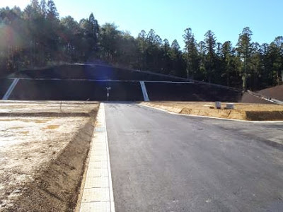 森林に囲まれた造成地の奥に、コンクリートの擁壁で土砂を抑えた構造があり、区画された造営地の中央に舗装済みの道路が整備された写真